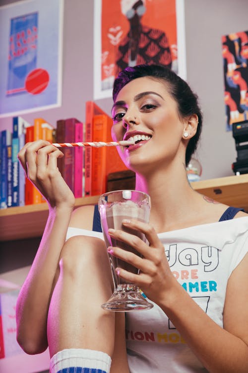 Low angle cheerful young lady in casual wear drinking sweet chocolate milkshake by using straw while spending time at home and looking at camera with smile