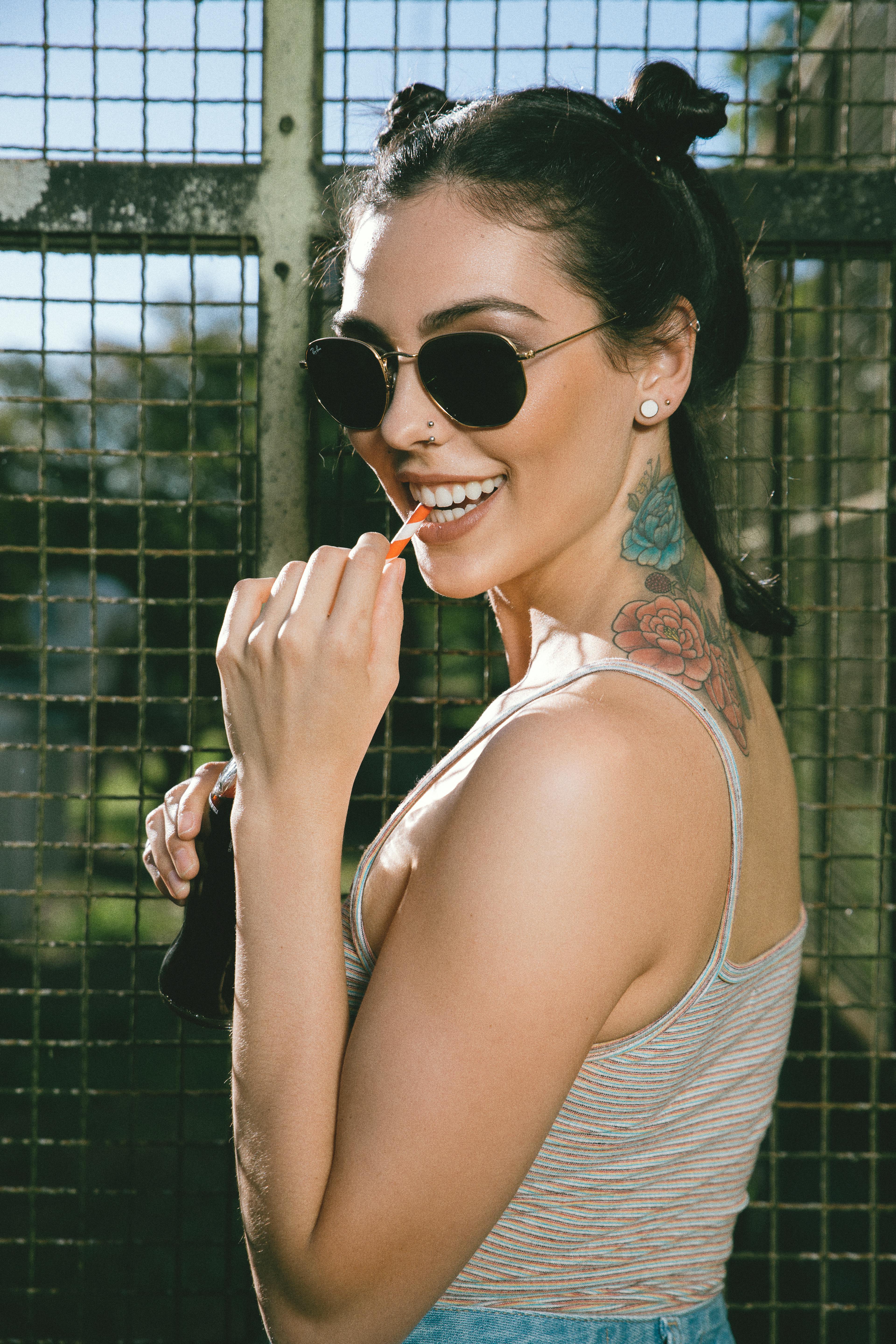 Young female with dark hair and tattoo on neck wearing summer top and sunglasses smiling with lollipop in mouth