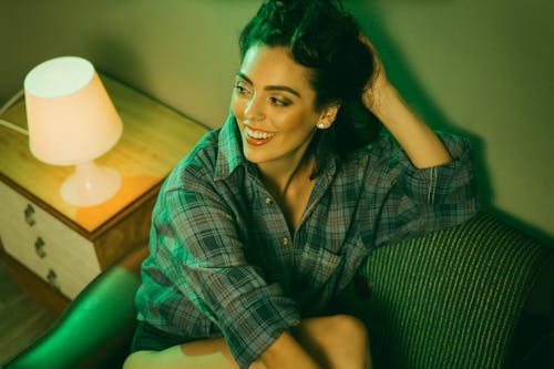 Smiling young female with dark hair in checkered shirt resting on couch next to nightlight in cozy room