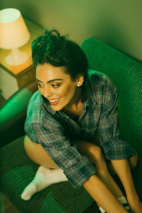 From above of joyful female with dark hair in checkered shirt and white socks sitting on green couch
