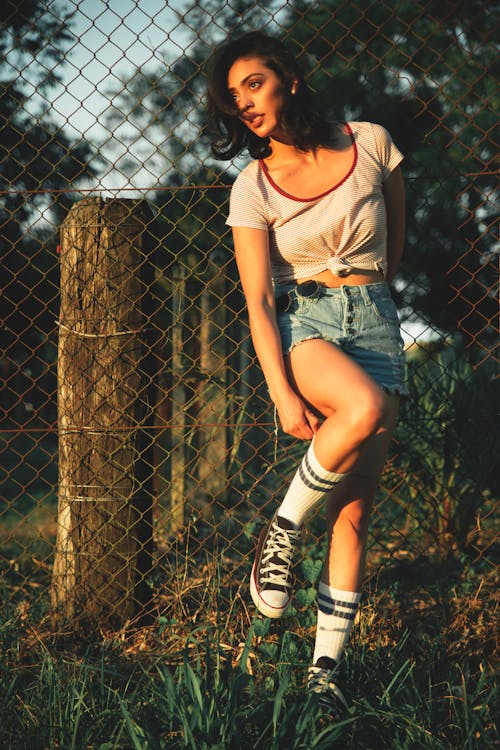 Full body of serious young ethnic female in t shirt and denim skirt and gaiters with sneakers standing against metal fence