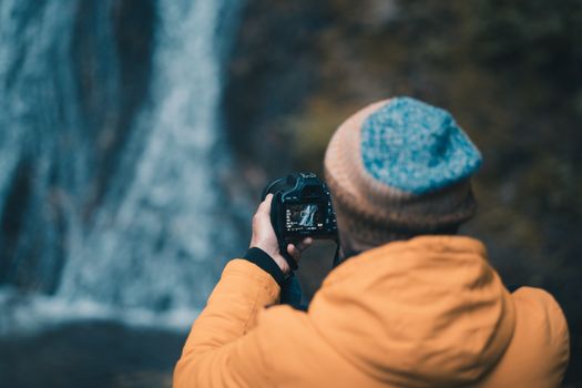 A photographer in a jacket and cap captures a waterfall in nature.