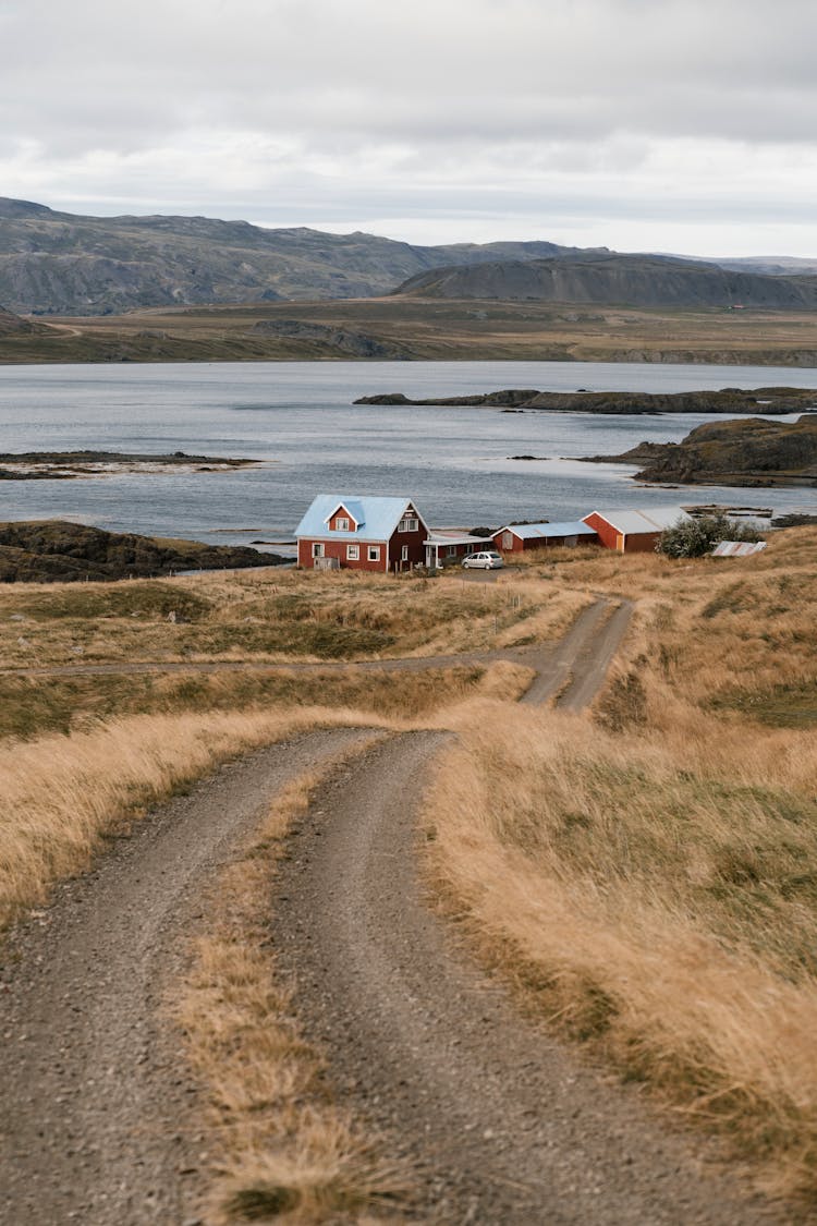 Country Road To Farm House Placed On Coastline With Hilly Cliffs Above Lake