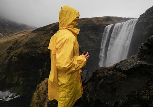 Side view of unrecognizable traveler in yellow raincoat standing on mountain close to high cascade in foggy weather in evening