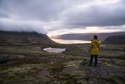 Back view of anonymous male traveler in raincoat standing in front of sea between ridge under cloudy sky in foggy weather