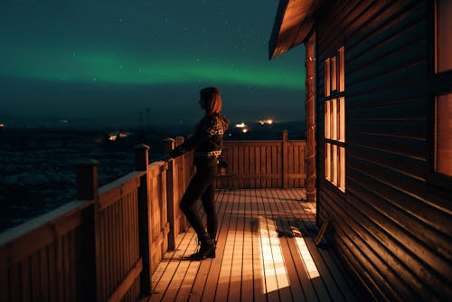 Side view of anonymous female in casual wear standing on wooden balcony of house and looking at majestic polar lights in dark sky