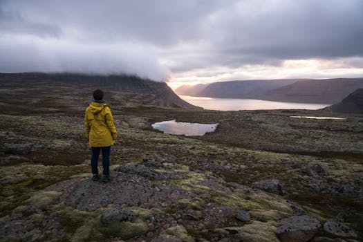 Back view of faceless traveler in yellow raincoat standing on rocky surface in front of sea between ridge covered with mist under cloudy sky at sunset