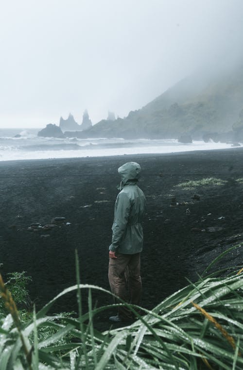 Side view of anonymous traveler in raincoat standing in front of stormy sea surrounded by ridge and grass covered with dew in foggy weather