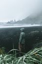 Side view of anonymous traveler in raincoat standing in front of stormy sea surrounded by ridge and grass covered with dew in foggy weather