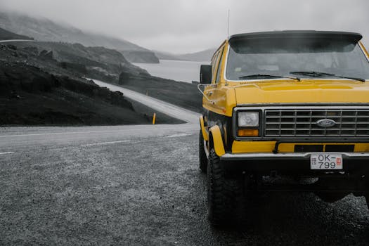 Yellow car parked on wet asphalt road passing between rough dark ridge and river under cloudy sky in foggy weather
