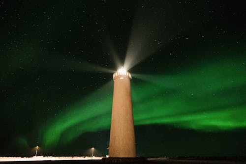 From below of high lighthouse tower burning in dark night against black sky with glowing green aurora light in north