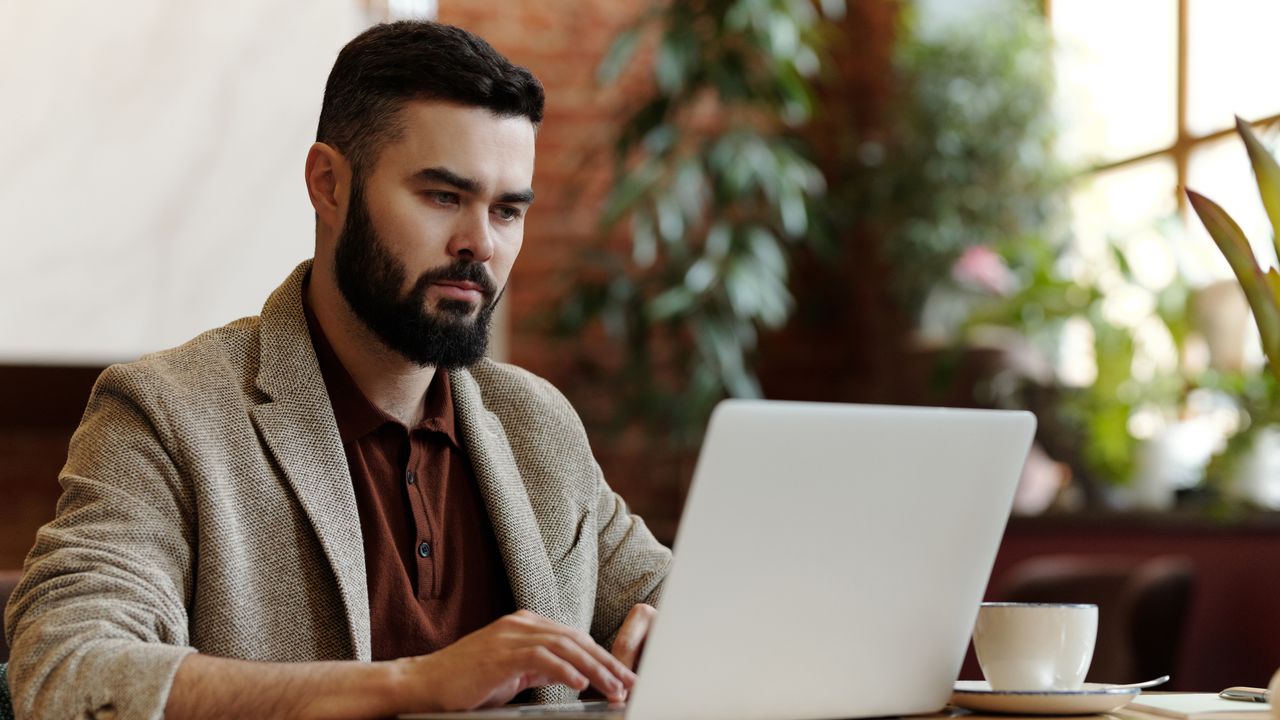 Businessman working on laptop in a cafe setting, surrounded by plants, sipping coffee.