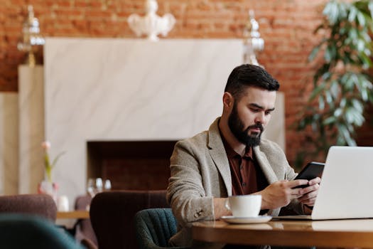 Bearded man using smartphone and laptop for remote work in a trendy coffee shop setting.