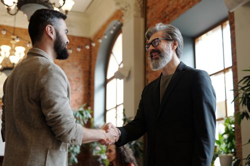 Free Two businessmen smiling during a handshake in a modern office setting, symbolizing success and partnership. Stock Photo