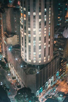 Aerial view of a modern skyscraper brightly lit at night in a bustling urban cityscape.
