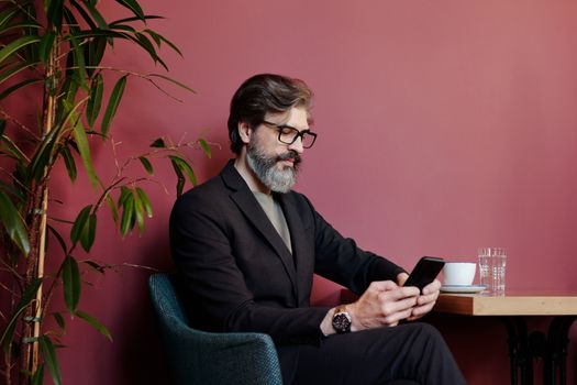 一个留着胡子、戴着眼镜的时髦人士正在现代咖啡馆里使用智能手机