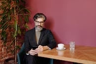 一位留着胡子的时尚男士坐在咖啡馆里，用智能手机喝咖啡