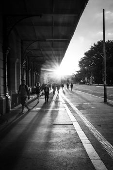 Black and white people walking through street under long roof of building near road in sunlight