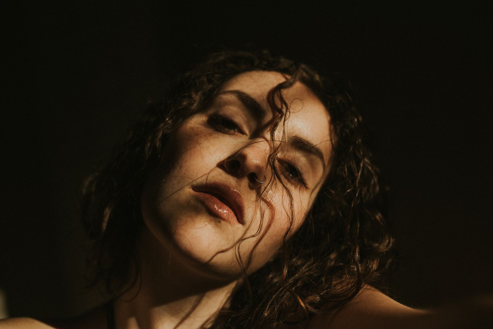 Woman with dark curly hair lit by warm side light against a dark background, looking toward the camera
