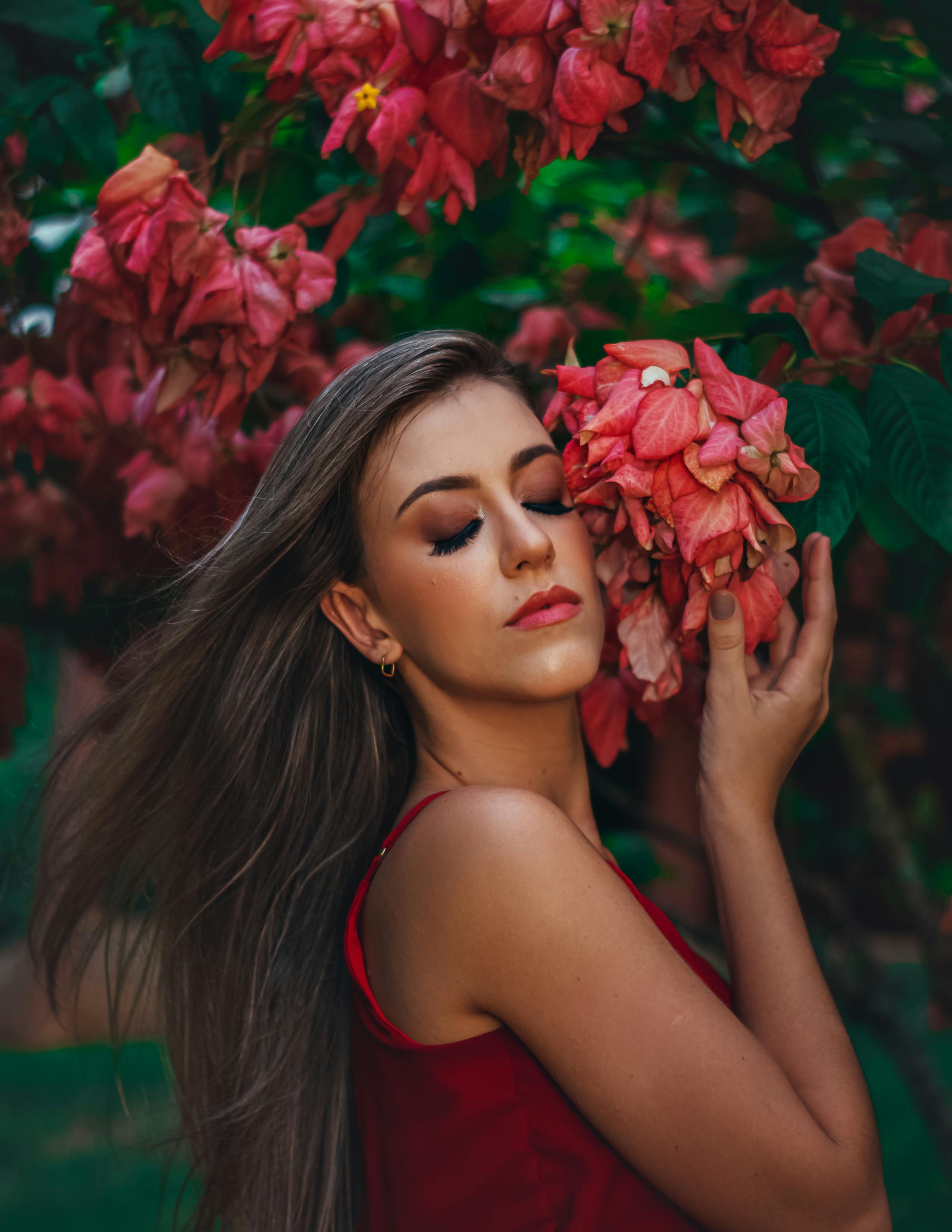 A beautiful woman closing her eyes and gently holding pink bougainvillea flowers in a serene outdoor setting.