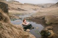Side view of young red haired female leaning on big stone in pond with transparent water formed of narrow river flow passing between mounts covered with sand in mist