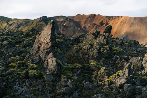 Spectacular view of sandy ridge in front of pointed rocks with rough partially mossy surface under cloudy sky in afternoon