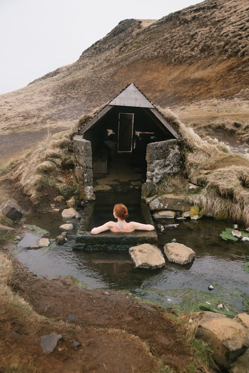Free Back view of anonymous red haired lady sitting in stone bath near small house and pond with pure water surrounded by sandy mountain under cloudy sky Stock Photo