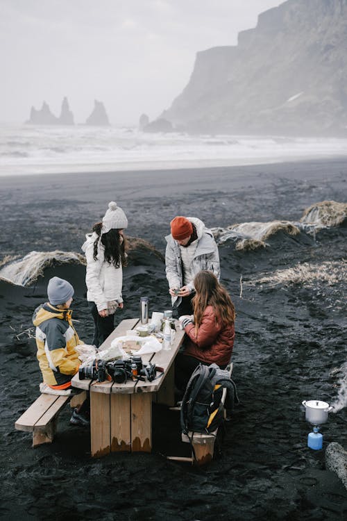 Free Group of people in outerwear sitting on wooden bench at table while preparing food on coast near ocean with waves forming foam and mountains under cloudy sky Stock Photo