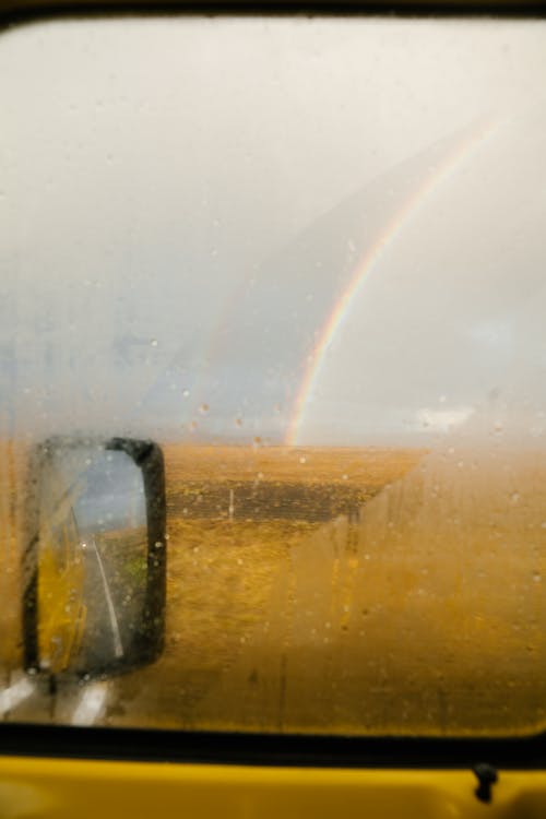 Yellow car with fogged up window parked on asphalt road and colorful rainbow on golden field in autumn under cloudy sky in countryside