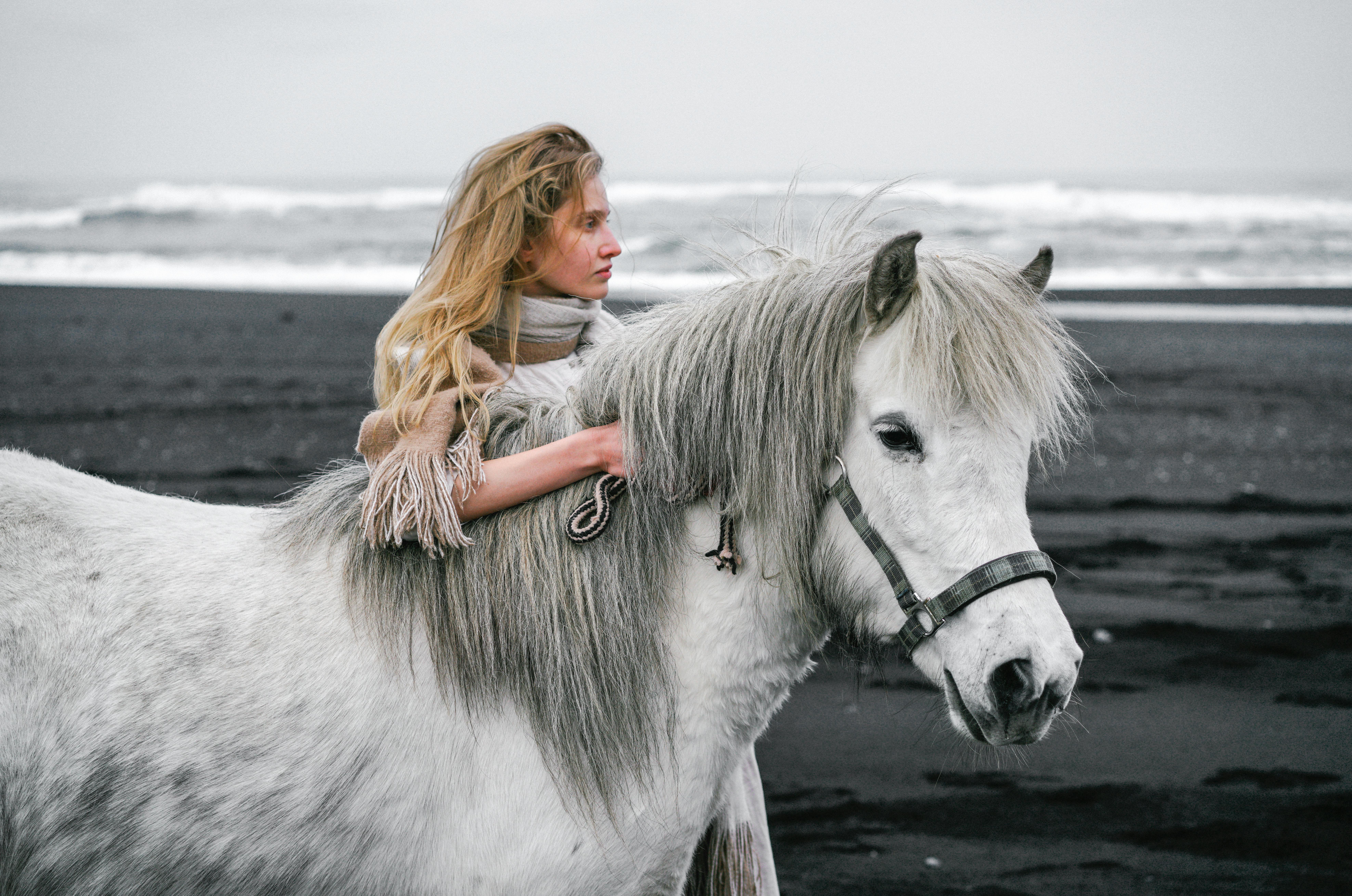 Side view of blond female palming mane of grey mare in bridle while standing on coast near sea with high waves in stormy weather and looking away