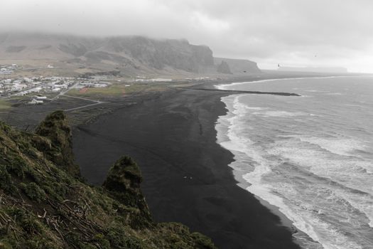 Picturesque scenery from cliff of foamy sea waves rolling over coast of ocean with black sand and village in mountains under 