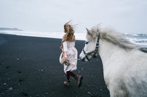 Back view of anonymous female wearing long white dress with scarf and warm boots walking along waving ocean on black sand shore together with white Icelandic horse