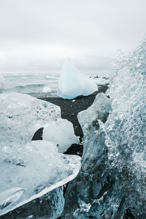 Free Amazing transparent clear ice pieces placed on black beach ground near calm ocean waves rolling over coast under gray cloudy sky in cold daytime Stock Photo