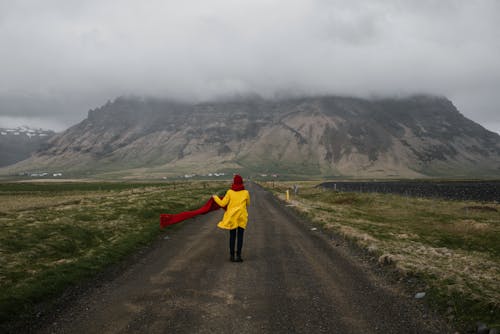 Free Back view of anonymous female wearing yellow coat and red scarf walking along empty road and admiring spectacular view of mountain with top hiding in fog in countryside during cold windy day Stock Photo