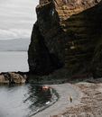From above of anonymous man walking along sandy beach along sea to rubber boat located near giant rock cliff on coast in sunny day