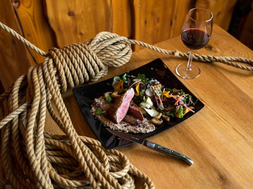From above of ceramic plate with beef steak pieces topped on mushroom sauce and vegetable salad served with knife and glass of red wine placed on wooden table with rope tied in knot