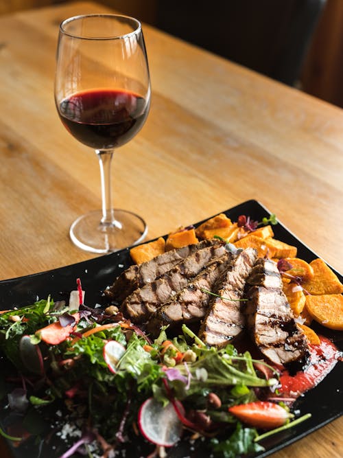 From above of glass of red wine and meat steak pieces served with tomato sauce and potato wedges with vegetable salad placed on ceramic plate on wooden table