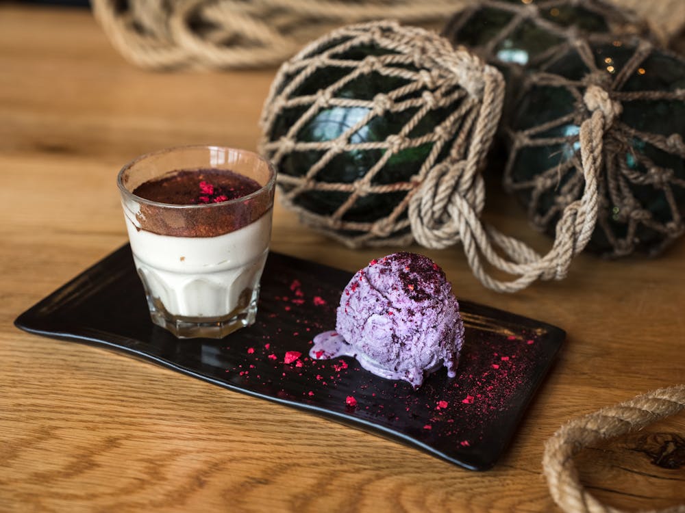 From above of glass of cream dessert with chocolate top served with blueberry ice cream scoop dusted with pink dye placed on ceramic plate on wooden table near black glass balls in grid of rope