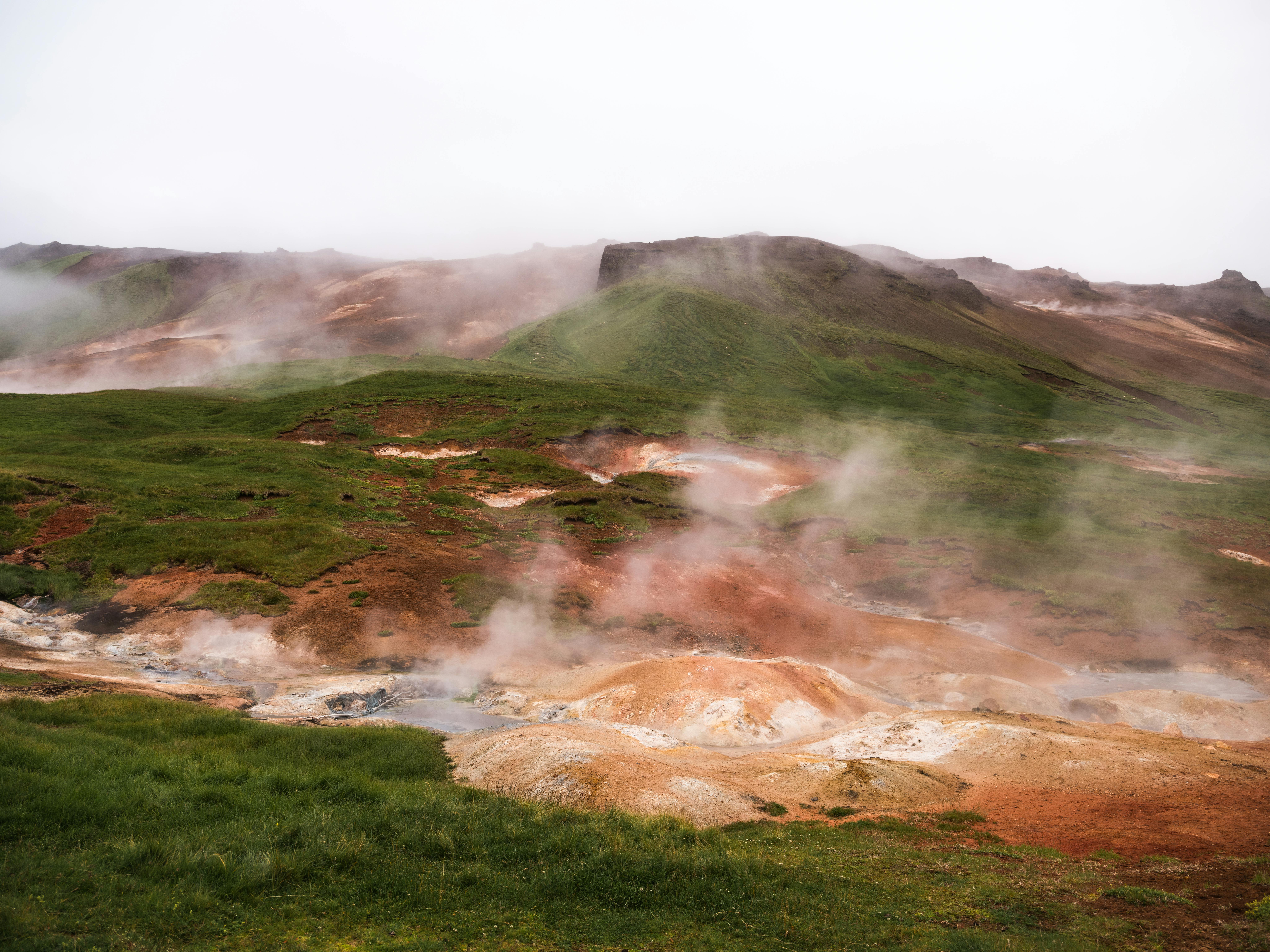 Hilled country with geysers blowing steam off among grass area against volcanic mountains in cloudy day
