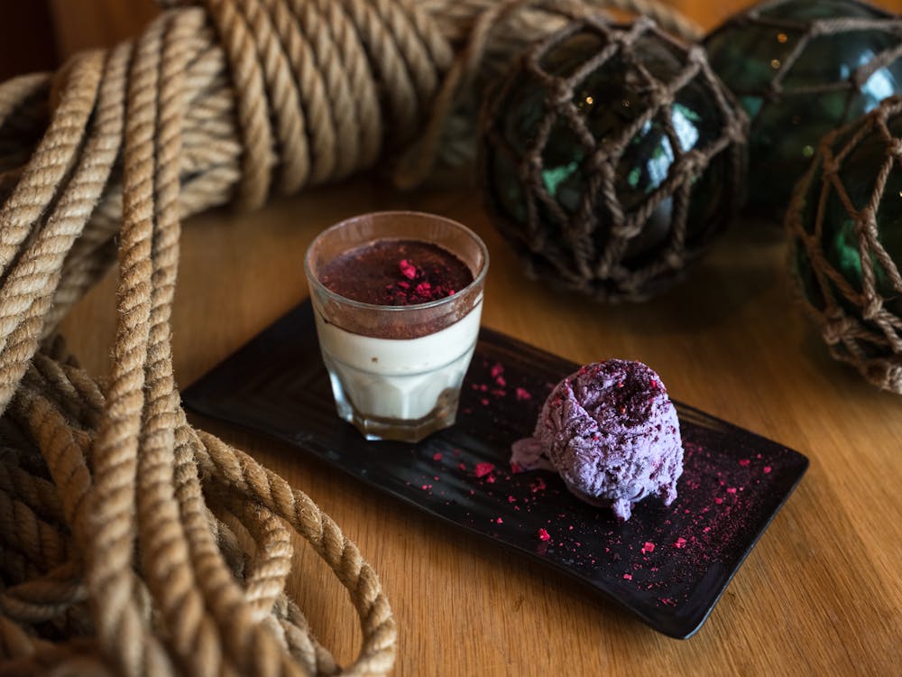 From above of glass of appetizing cream dessert with chocolate top served with blueberry ice cream garnished with pink dye on ceramic plate surrounded by rope tied in knot and dark green glass balls in grid