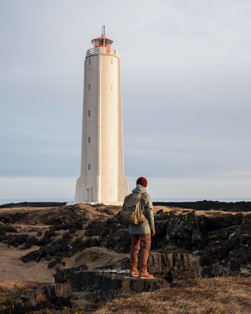 Free Back view of full length unrecognizable male traveler in outerwear and backpack standing on rock on grass terrain and observing lighthouse in cloudy evening Stock Photo