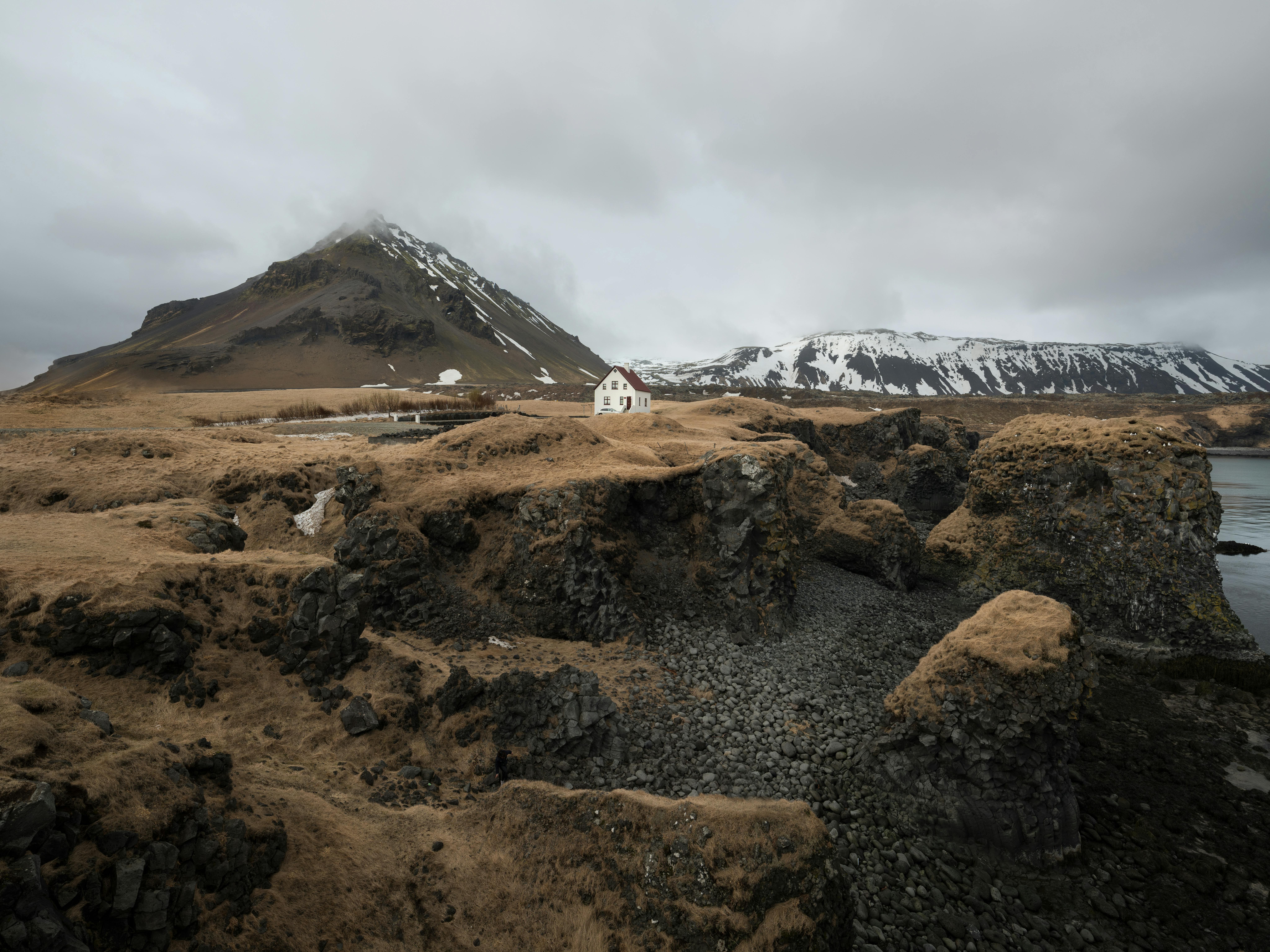 Picturesque view of rocky autumn seashore with snowy mountains and remote rural house under cloudy gray sky