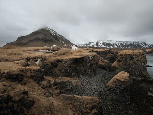 Free Picturesque view of rocky autumn seashore with snowy mountains and remote rural house under cloudy gray sky Stock Photo