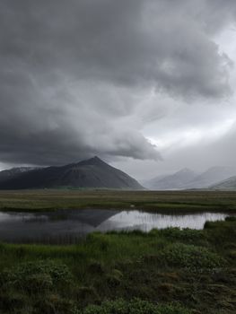Majestic view of thick dark gray clouds floating over calm lake surrounded with mountains and green grass