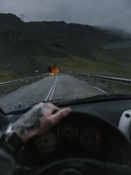 Crop unrecognizable person with tattooed hand o steering wheel driving into tunnel on mountainous road near rocky formations on cloudy evening
