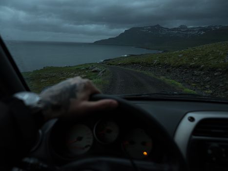 Crop anonymous person with hand on steering wheel driving along narrow rural road towards calm sea and dramatic rocky mountains under cloudy dark sky at night