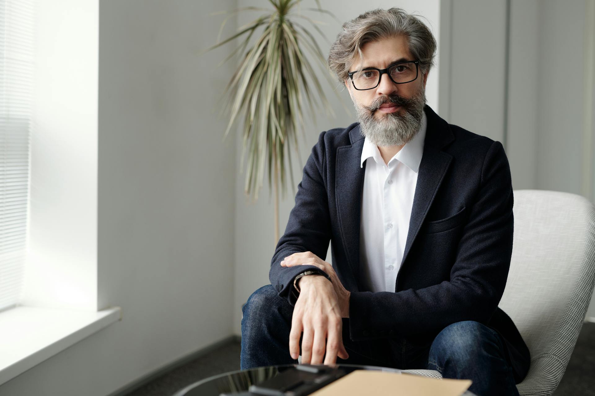 A mature bearded businessman in a suit sitting and looking confidently at the camera indoors.