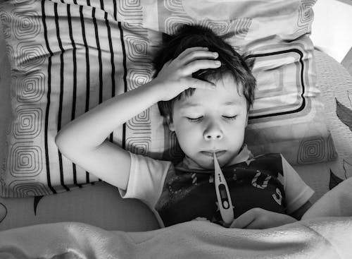 Free Black and white overhead view of child in casual clothing measuring body temperature with electronic thermometer while lying on bed under blanket Stock Photo