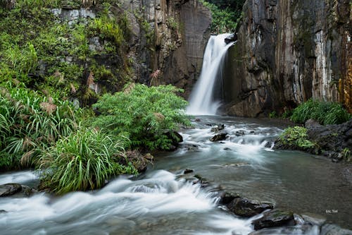 Long exposure of high waterfall of broad mountain river surrounded by steep rocks and greenery in ravine