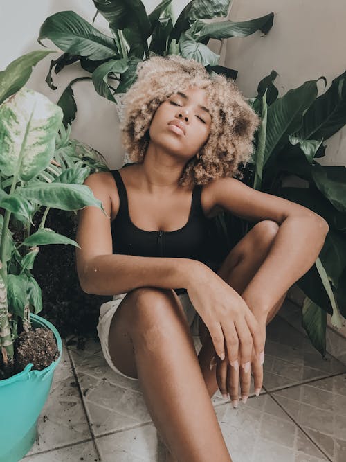 Alluring ethnic woman in casual clothing with curly hair resting on tile paved floor in room with eyes closed surrounded by pot plants